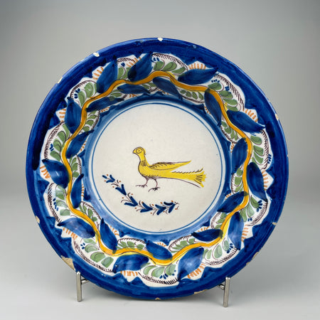 Antique Spanish Manises ceramic plate with hand-painted yellow bird, polychrome floral border, and scalloped blue rim, photographed upright to highlight its 27 cm width and folk art style for rustic Mediterranean wall decor or collector display.