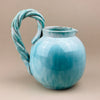 Blue ceramic pitcher with a twisted handle on a beige background. Side profile of the vintage Belgian jug attributed to Alexandre de Wemmel, showing the balanced proportions, rounded body, and spout detail. The turquoise glaze highlights the studio-crafted form, representative of 1950s mid-century European ceramics.