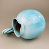 Light blue ceramic vase with a textured surface on a beige background. Rear view of the jug highlighting the smooth curves and glaze pooling near the base. The twisted handle emerges as a focal point, enhancing its sculptural quality and making it a statement piece for art pottery collectors.