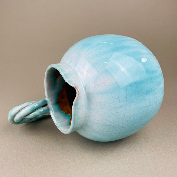 Light blue ceramic vase with a textured surface on a beige background. Rear view of the jug highlighting the smooth curves and glaze pooling near the base. The twisted handle emerges as a focal point, enhancing its sculptural quality and making it a statement piece for art pottery collectors.