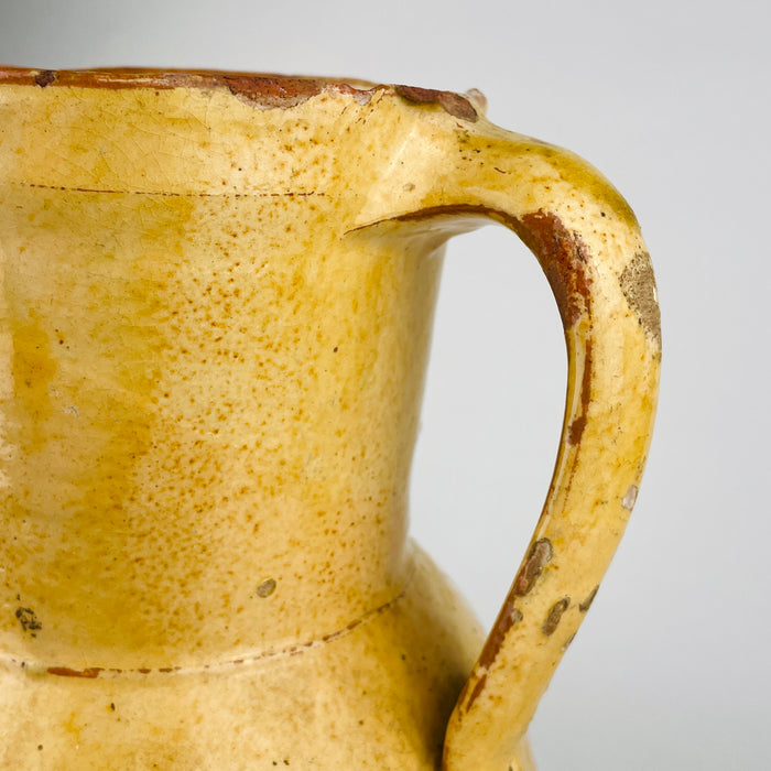 Antique Spanish jug with handle in yellow earthenware glaze, combining a strong practical form with weathered surface texture. The old pottery body shows losses around the mouth, rubbed glaze on the handle, and exposed red clay at the base, all of which add to its decorative appeal in Mediterranean decor, farmhouse decor, and wabi sabi decor.