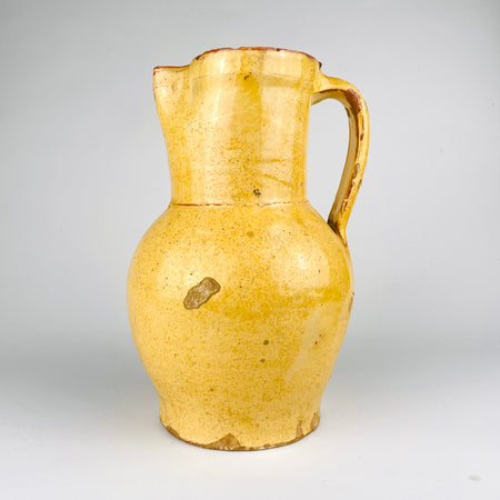 Nineteenth century Spanish terracotta jug with ochre yellow glaze, showing the rounded profile and narrow upper section typical of rural earthenware pottery. The vessel has surface losses and worn edges that reveal the red clay body beneath, giving it strong antique character for Mediterranean decor and antique kitchen pottery displays.