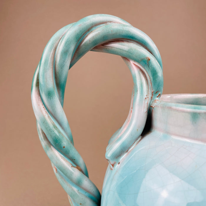 Close-up of a ceramic pitcher with a twisted handle on a beige background. Focus on the twisted rope handle, demonstrating the sculptural craftsmanship of Alexandre de Wemmel. This bold handle is a recurring motif in his mid-century ceramics, celebrated for merging utility with strong artistic expression.