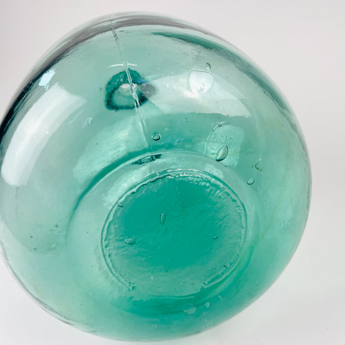 Vintage glass damajuana from Spain with a blue demijohn vase form, rounded body, long neck, and raised V. Ayelense lettering. The aqua glass color and visible production marks support its identity as a wine carboy, while its decorative scale works well in Mediterranean decor, coastal decor, farmhouse decor, and vintage home decor.
