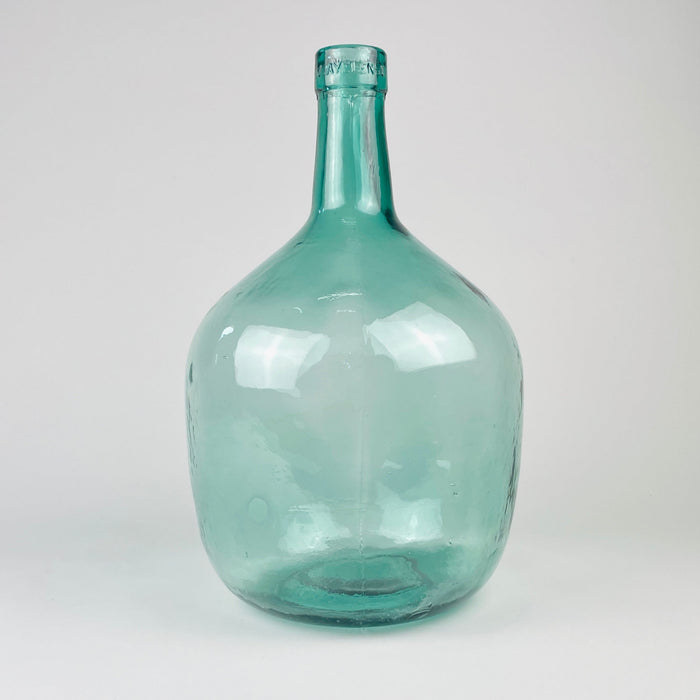 V. Ayelense glass demijohn from Spain, made in blue green aqua glass with a round body, thick neck rim, visible bubbles, and raised lettering identifying the maker. The piece functions today as a demijohn vase, wine carboy display bottle, or sculptural glass accent for farmhouse decor, coastal decor, and vintage kitchen styling.
