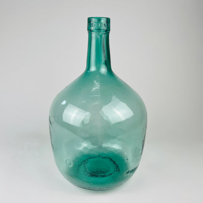 Large blue glass carboy with Spanish demijohn form, rounded storage bottle body, and narrow neck with embossed maker lettering. Originally intended for wine or liquid storage, this vintage glass vessel now works as a decorative vase for tall stems, dried flowers, branches, or empty display in Mediterranean decor and rustic home decor.