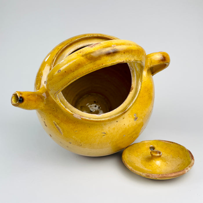 Ochre yellow French terracotta pottery water jug with the form of an antique gargoulette pot. The vessel includes a lid with small air hole, a rounded knob, a short pouring spout, and carrying handles, while the worn glaze and exposed terracotta support use as a decorative ceramic centerpiece, pottery vase, or Mediterranean decor object.