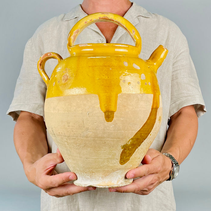 Antique French ceramic water jug in yellow glazed terracotta, with a rounded body, side spout, arched handle, loop handle, and removable lid. The pottery surface shows historic wear, glaze chips, scratches, exposed clay, and base abrasions, making it an expressive decorative vessel for antique pottery collectors and vintage home decor.