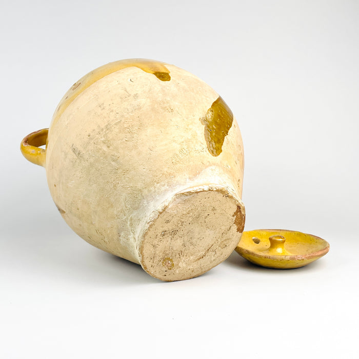 Antique French water jug with lid, made from yellow glazed terracotta and shaped as a large gargoulette pot. The warm glaze, raw clay lower body, arched handle, spout, side loop handle, and visible wear identify it as a utilitarian pottery vessel, now well suited for French country decor, vintage kitchen display, and rustic shelf styling.