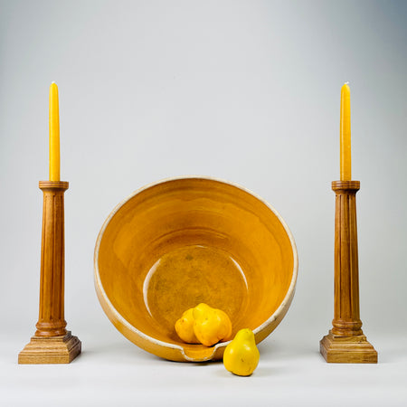 This image shows a large antique French terracotta bowl with a warm amber ochre glaze, displayed upright to highlight its wide 40.5 cm form and deep interior. The glaze shows natural variation, subtle texture that reflect hand thrown craftsmanship typical of late 19th century French farmhouse pottery. This bowl suits farmhouse decor, French country decor, and Rustic Home as a sculptural centrepiece or display vessel. The bowl is styled alongside wooden candleholders.
