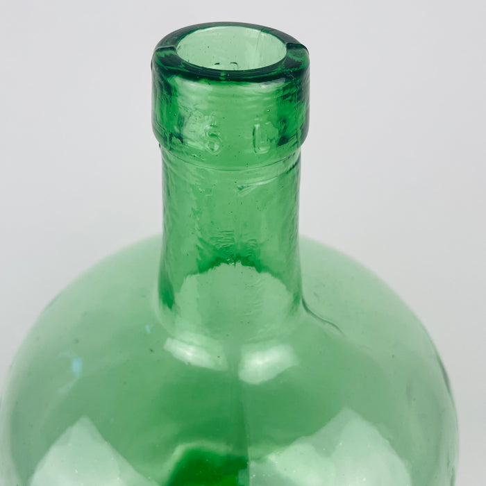 Embossed VIRESA maker name on the reinforced neck of a vintage Spanish green glass demijohn from Benigànim, Valencia, circa 1960s. The mould blown construction produces a visible seam, gentle waviness and trapped air bubbles that document its industrial manufacture. This marked 5 L wine carboy now works as authentic Spanish vintage decor.