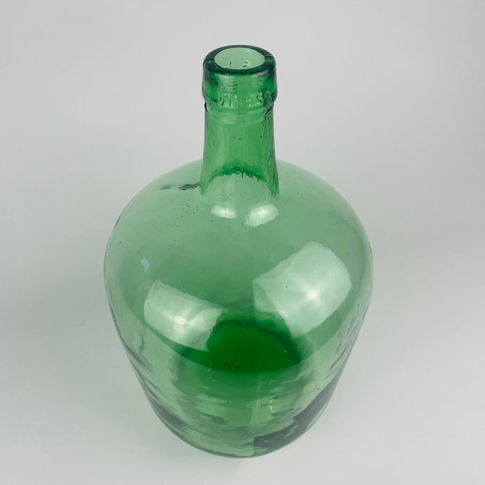 Thick rounded mouth and VIRESA lettering on a vintage 5 L Spanish demijohn vase made in Benigànim, Valencia, circa 1960s. The green mould blown glass shifts from pale aqua across the neck to deep emerald at the reinforced rim, while tiny bubbles, ripples and irregularities animate the surface. A practical wine vessel repurposed for vintage glass decor.