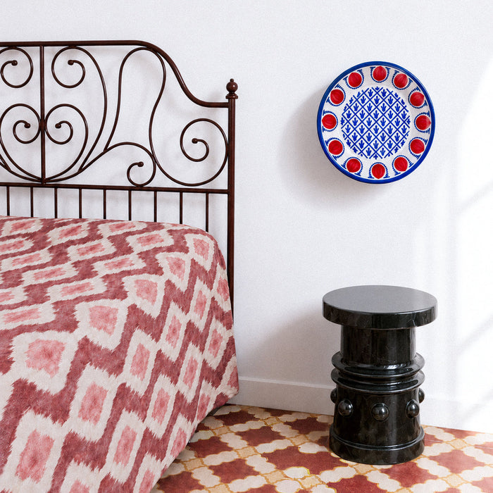 A styled bedroom corner featuring a bed with a pink and red ikat patterned duvet, a dark metal headboard, a black cylindrical side stool, and a decorative blue and red spanish fajalauza ceramic wall plate on a white wall. Granada pottery bowl in the Fajalauza tradition, hand painted with red pomegranates, dotted blue arches, and a blue geometric lattice filled with foliate brush marks. This contemporary Spanish ceramic bowl works as a fruit bowl, table centerpiece, or wall bowl for Mediterranean decor and r